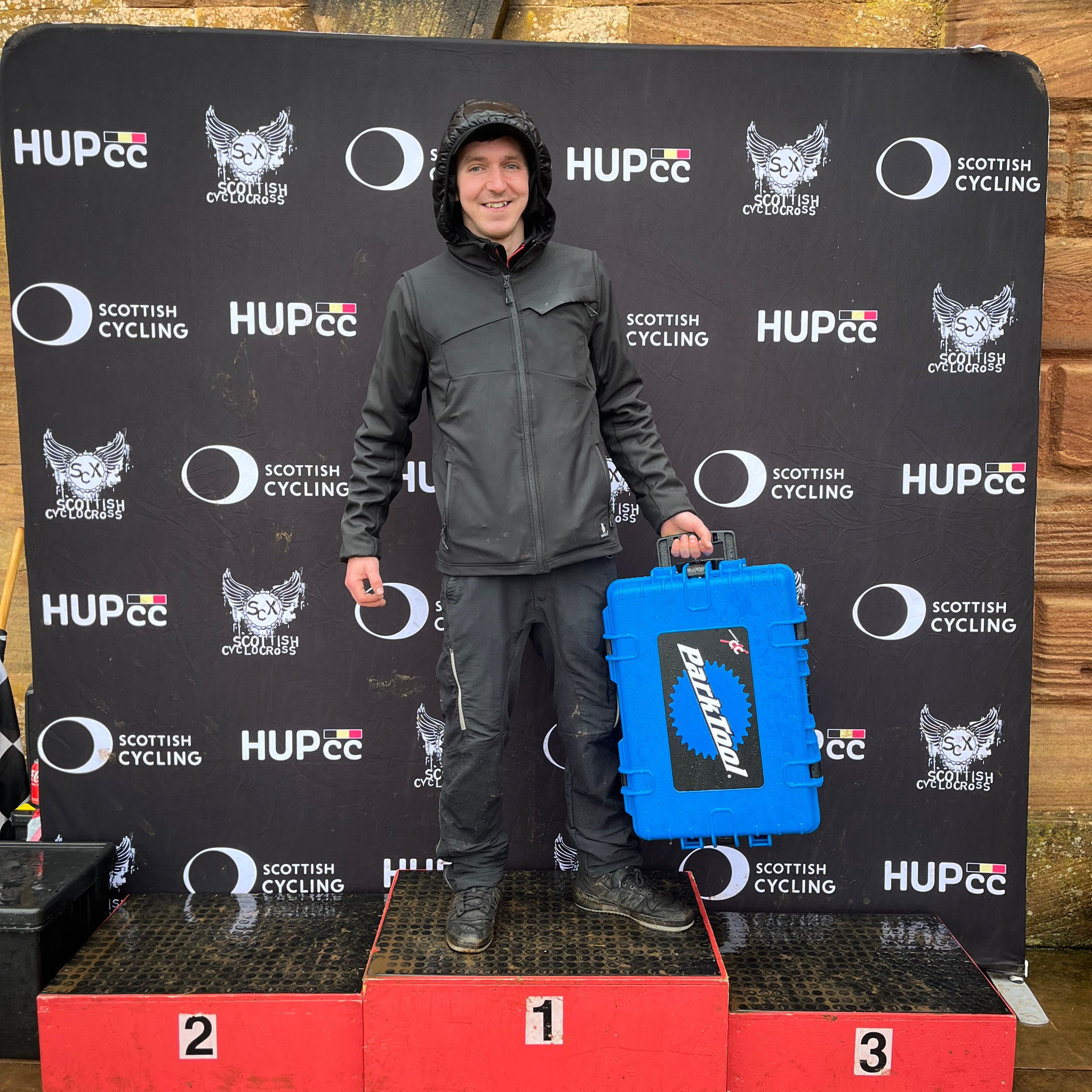 A man in a dark gray jacket and black pants stands on a red three-step podium holding a bright blue Park Tool hard case in front of a black HUP26 cycling backdrop.